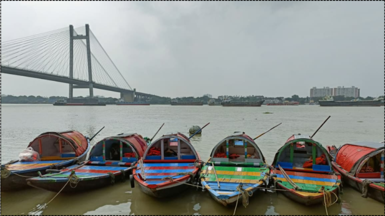 Six colorful traditional wooden boats moored on the Hooghly River in Kolkata, with the Vidyasagar Setu cable-stayed bridge spanning the water under an overcast sky.